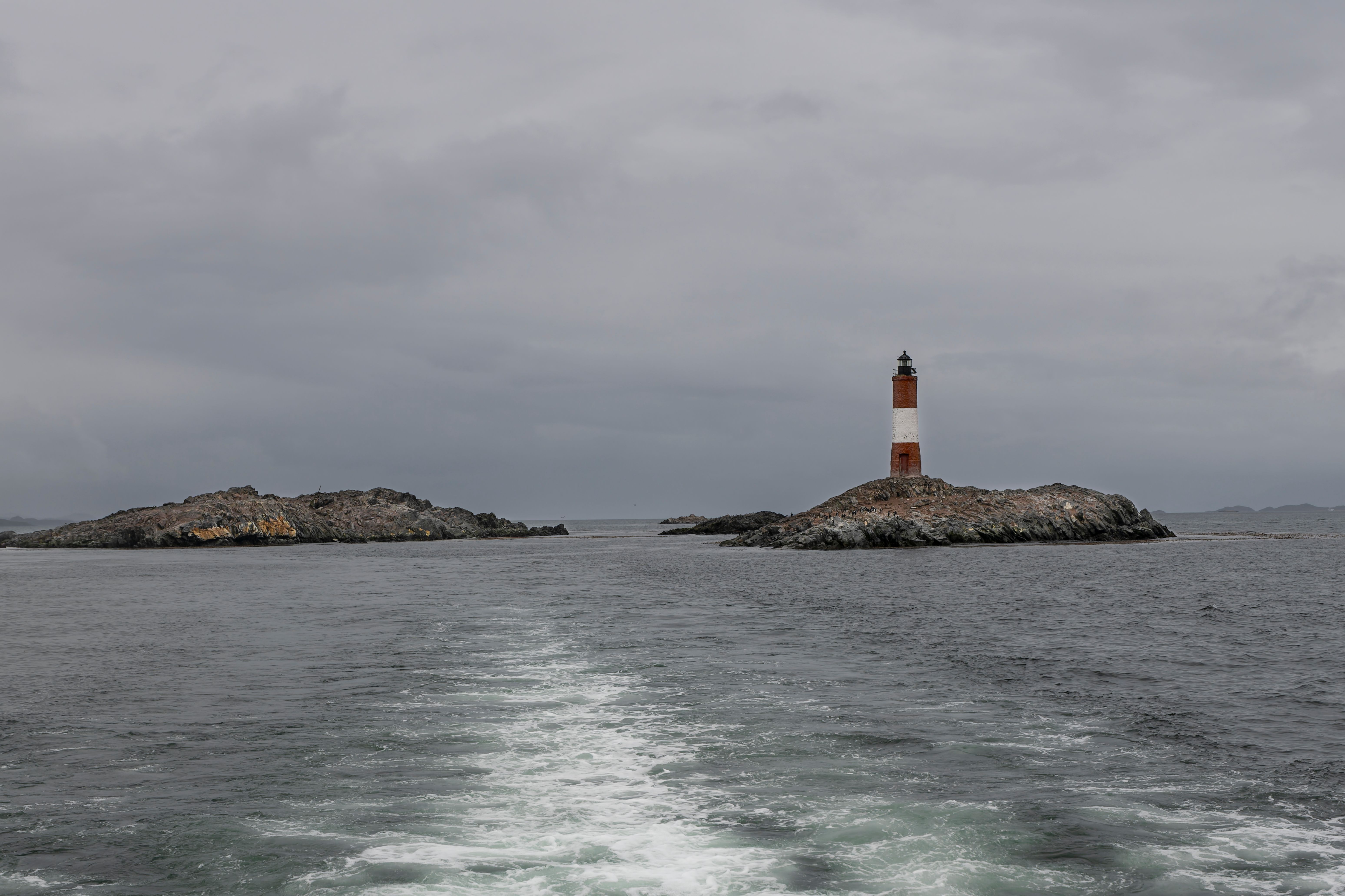 Red and white lighthouse stands on rocky island amidst calm sea under cloudy skies.